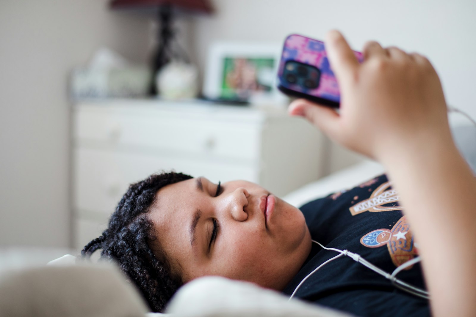 A young person lying on a bed at home, holding a phone above them.